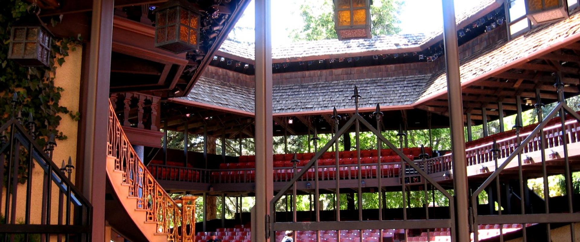 The image shows the interior of a wooden, open-air theater structure with tiered seating, a central stage area, and ornate lanterns hanging from the ceiling, viewed through a foreground gate.