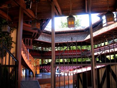 The image shows the interior of a wooden, open-air theater structure with tiered seating, a central stage area, and ornate lanterns hanging from the ceiling, viewed through a foreground gate.