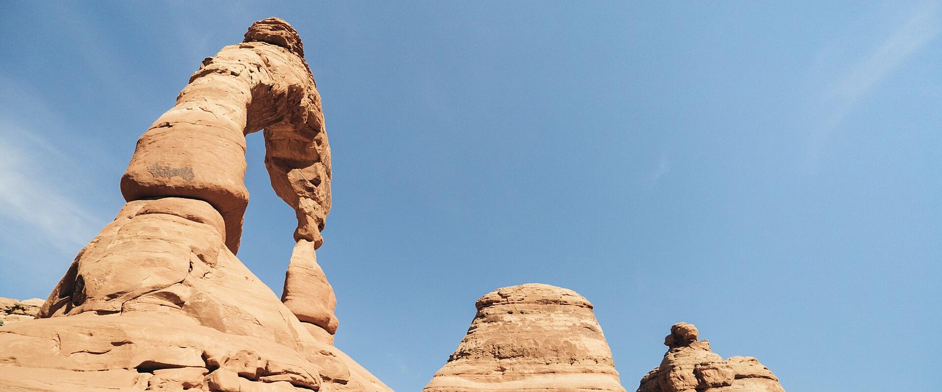 Low-angle view of a freestanding red sandstone arch against a blue sky, with a smaller rock formation visible to the right.