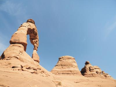 Low-angle view of a freestanding red sandstone arch against a blue sky, with a smaller rock formation visible to the right.