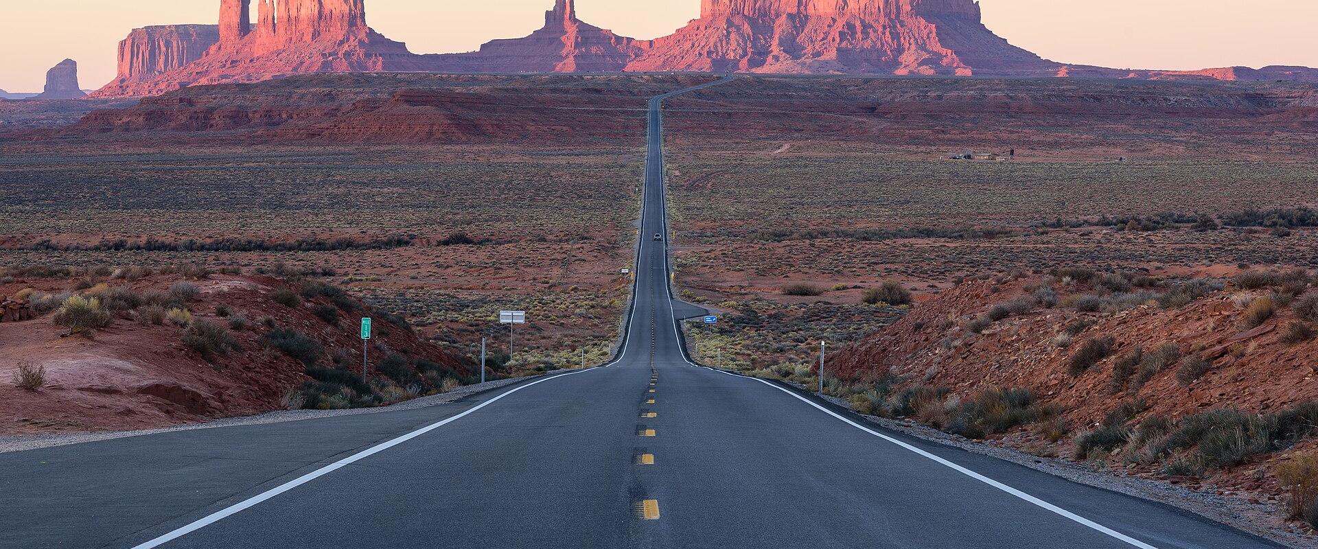 A view of a straight, paved highway stretching into the distance, flanked by desert scrub and the iconic red rock buttes of Monument Valley, with a green mile marker sign visible on the left shoulder.