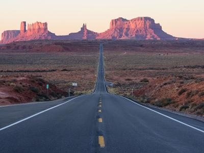 A view of a straight, paved highway stretching into the distance, flanked by desert scrub and the iconic red rock buttes of Monument Valley, with a green mile marker sign visible on the left shoulder.