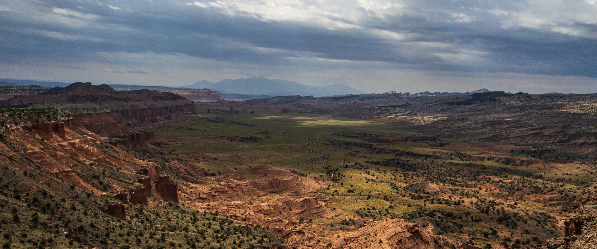 A wide landscape photograph showing a high vantage point looking down into a large, flat valley floor surrounded by steep, reddish-brown sandstone cliffs and mesas, with a dramatic, cloudy sky above.