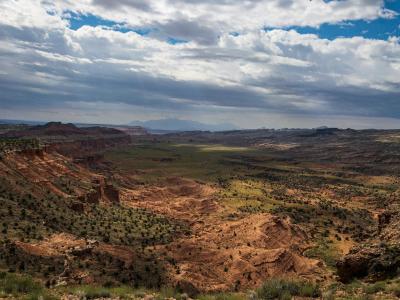 A wide landscape photograph showing a high vantage point looking down into a large, flat valley floor surrounded by steep, reddish-brown sandstone cliffs and mesas, with a dramatic, cloudy sky above.