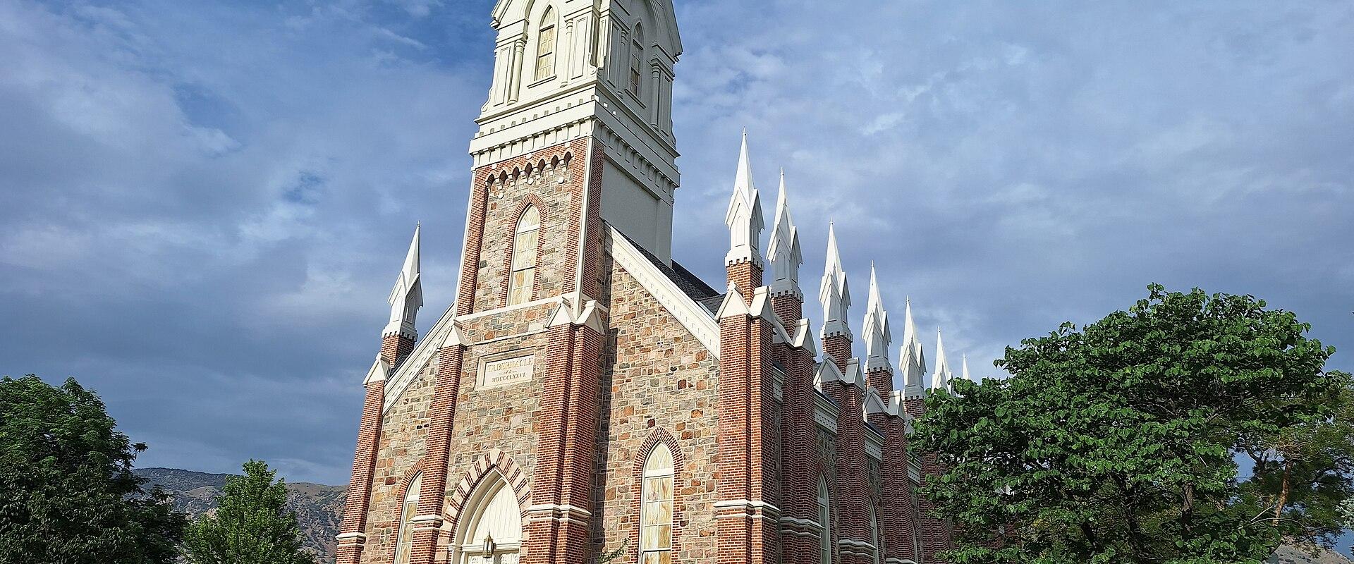 The image shows a large, ornate religious building with a central white steeple, red brick walls, and multiple smaller white spires along the roofline, set against a blue sky with trees in the foreground.