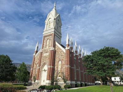 The image shows a large, ornate religious building with a central white steeple, red brick walls, and multiple smaller white spires along the roofline, set against a blue sky with trees in the foreground.