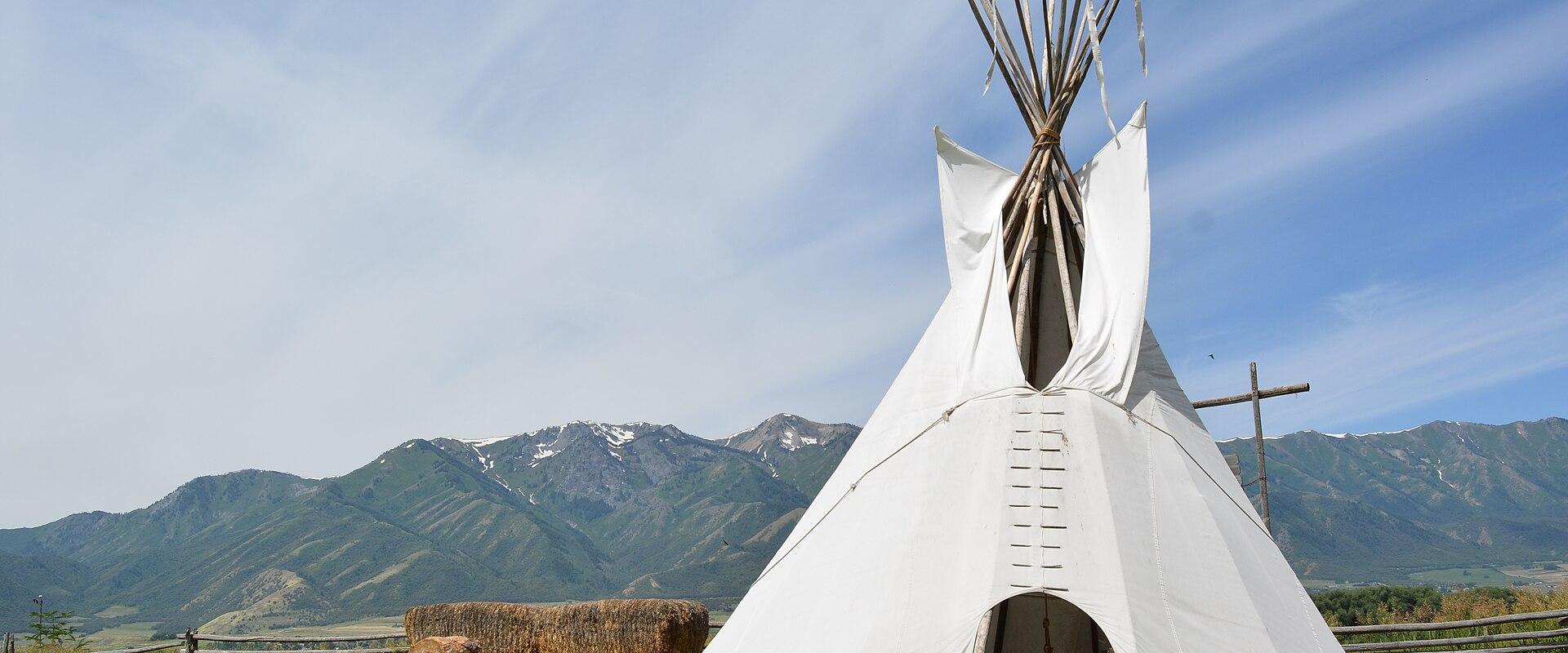 A white canvas teepee (tipi) situated on a grassy field, with a wooden fence, a bale of hay, and a mountain range in the background.