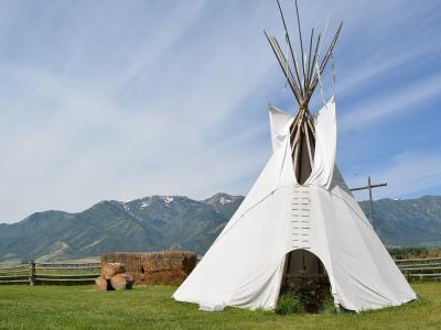 A white canvas teepee (tipi) situated on a grassy field, with a wooden fence, a bale of hay, and a mountain range in the background.