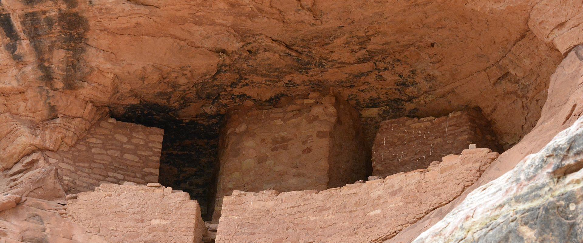 Ancestral Puebloan cliff dwelling ruins (masonry walls) built into the recess of a large, reddish sandstone alcove.