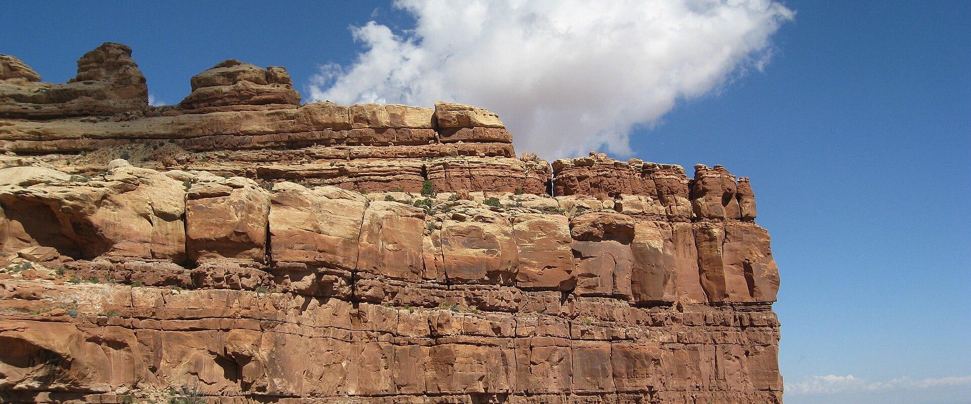 A view of a reddish-brown sandstone cliff face and mesa top against a blue sky, with a distant view of a flat valley floor in the background.