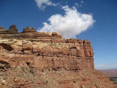 A view of a reddish-brown sandstone cliff face and mesa top against a blue sky, with a distant view of a flat valley floor in the background.