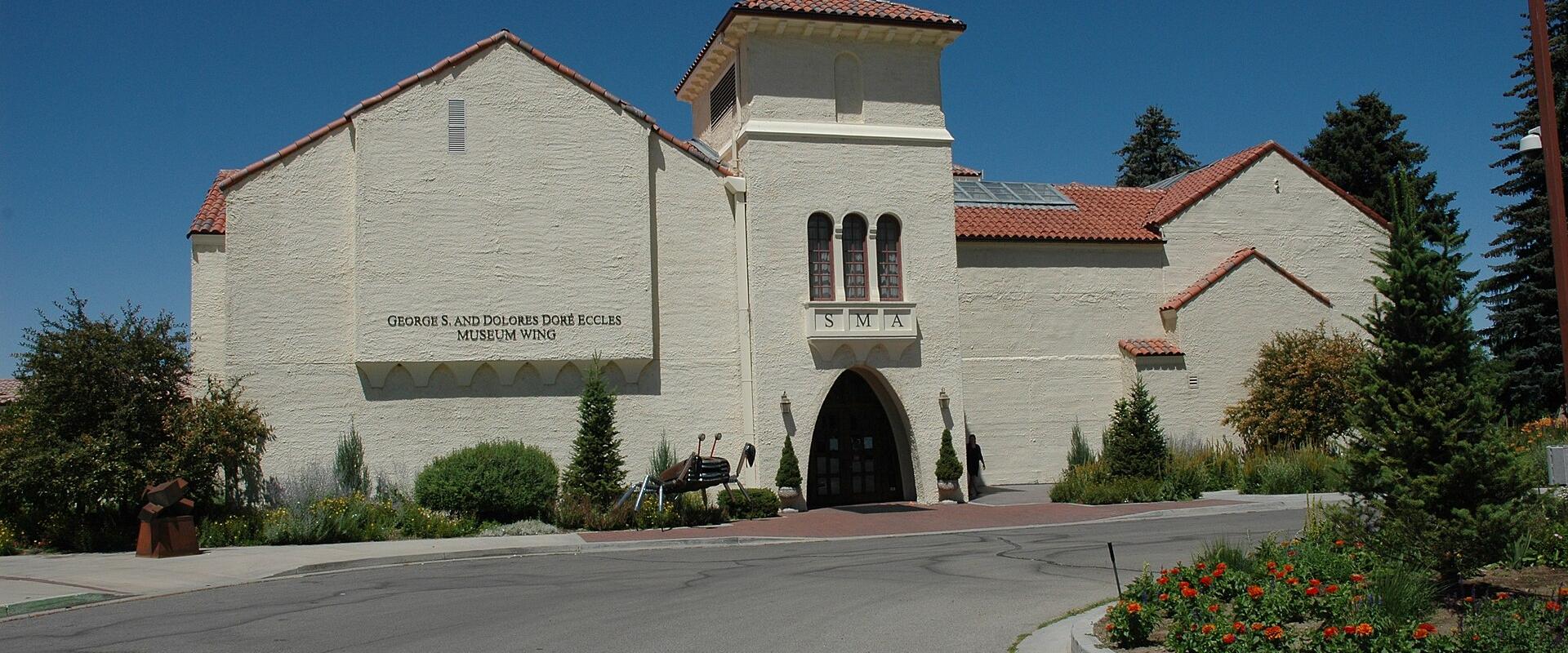 A cream-colored stucco building with a red tile roof, featuring signage that reads "GEORGE S. AND DOLORES DORE ECCLES MUSEUM WING" and "SMA" above an arched entrance.