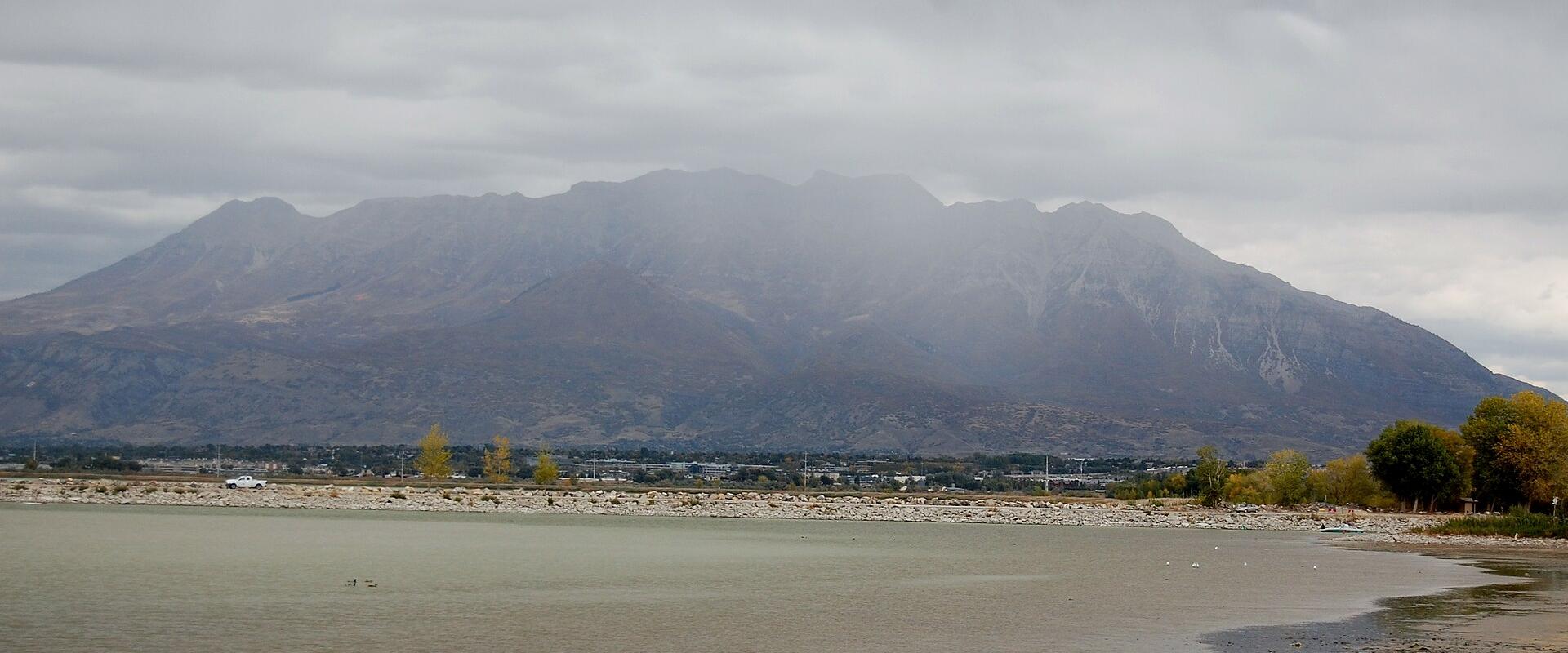 A wide landscape view showing a large body of water in the foreground, a rocky shoreline, a line of trees, and a large, rugged mountain range in the background under a cloudy sky.