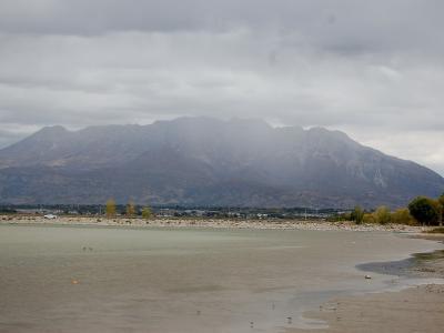 A wide landscape view showing a large body of water in the foreground, a rocky shoreline, a line of trees, and a large, rugged mountain range in the background under a cloudy sky.