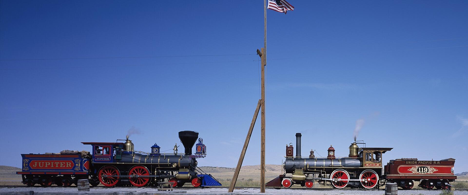 Two historic steam locomotives, the Jupiter and the No. 119, facing each other on parallel tracks with a flagpole between them in an open, arid landscape.
