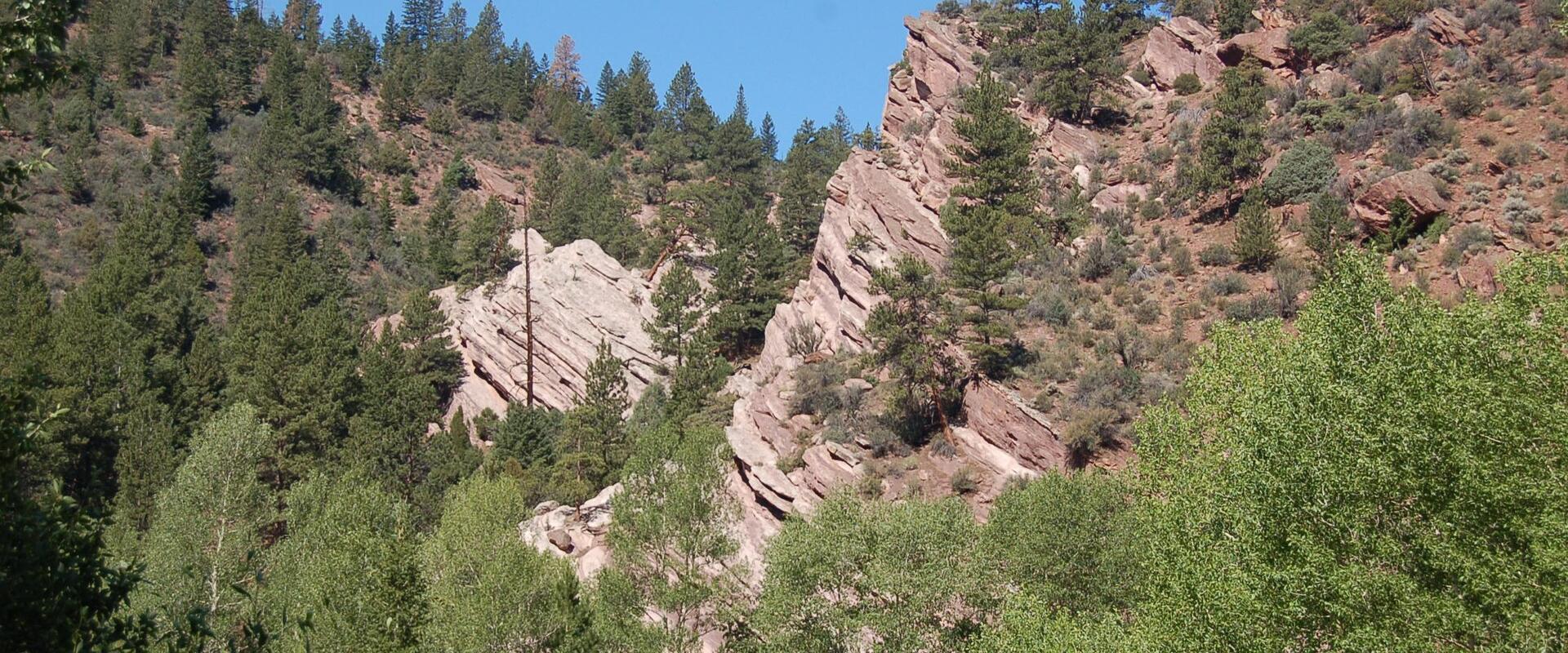 A view of a steep, rocky hillside with reddish-brown rock formations and green coniferous trees, featuring a road barrier in the foreground.