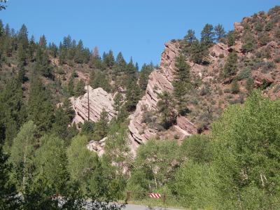 A view of a steep, rocky hillside with reddish-brown rock formations and green coniferous trees, featuring a road barrier in the foreground.