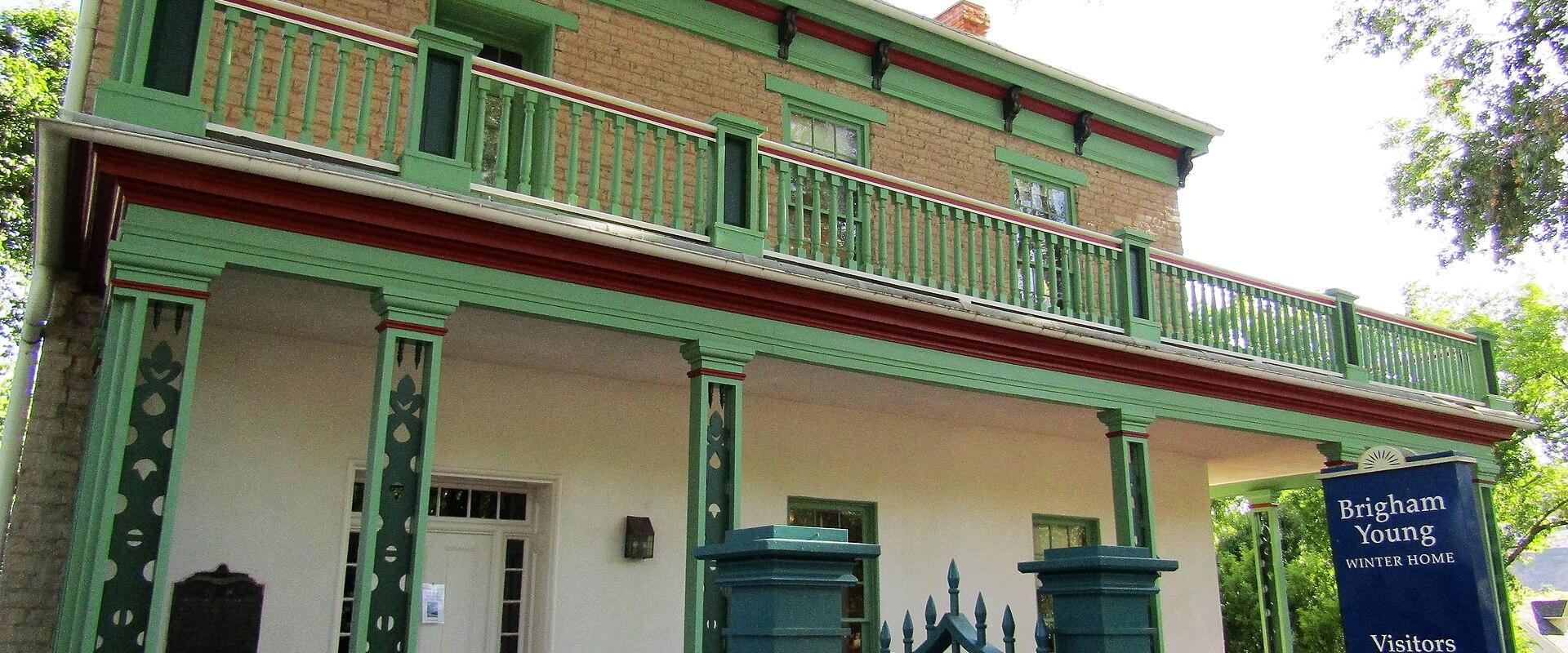 A two-story building with a green and white painted facade, a wraparound porch, and a prominent blue sign on the right that reads "Brigham Young Winter Home" along with "Visitors Welcome" and "Open Free Tours".