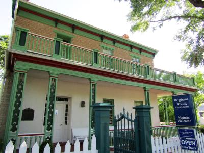 A two-story building with a green and white painted facade, a wraparound porch, and a prominent blue sign on the right that reads "Brigham Young Winter Home" along with "Visitors Welcome" and "Open Free Tours".