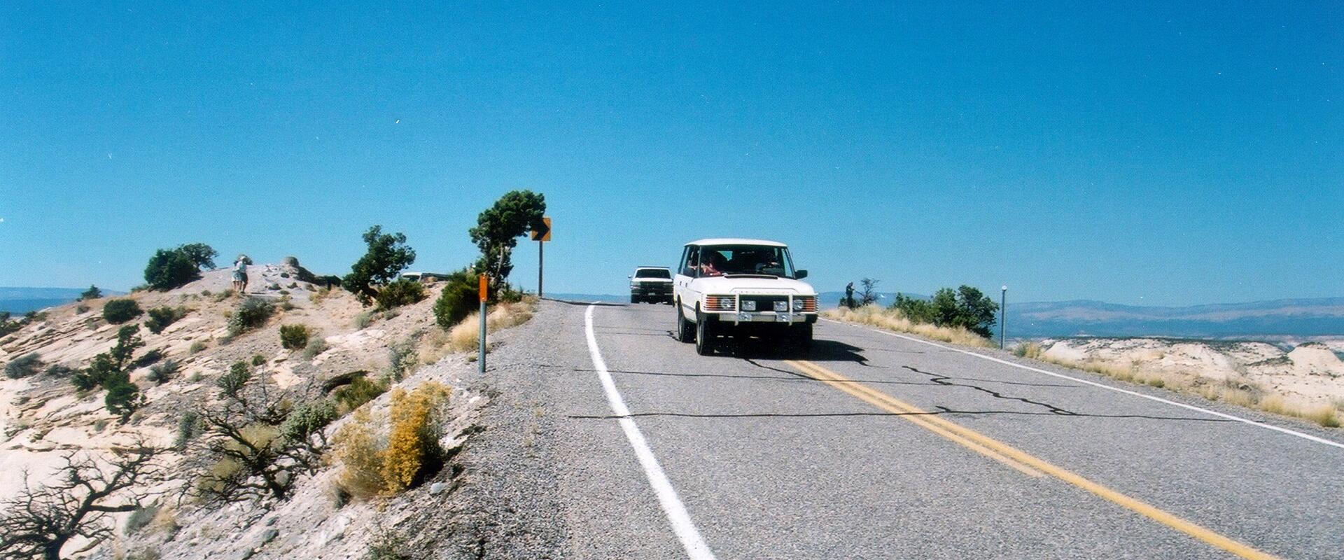 A white SUV and a dark vehicle are driving on a paved, two-lane road through an arid, desert landscape with sparse vegetation, set against a clear blue sky.