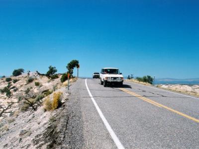 A white SUV and a dark vehicle are driving on a paved, two-lane road through an arid, desert landscape with sparse vegetation, set against a clear blue sky.