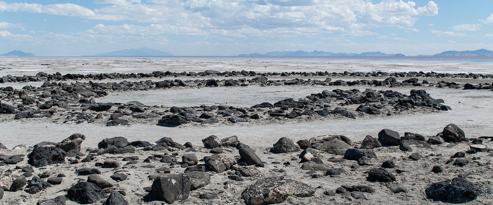 A landscape photograph showing a flat, white salt flat in the foreground with scattered dark rocks, leading to a body of water and distant mountains under a blue sky with clouds.