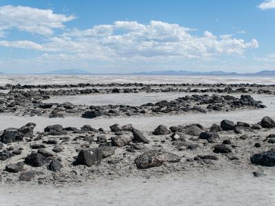 A landscape photograph showing a flat, white salt flat in the foreground with scattered dark rocks, leading to a body of water and distant mountains under a blue sky with clouds.
