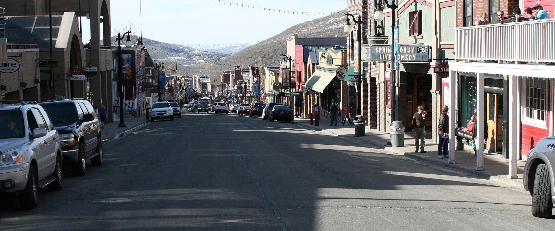 A paved main street lined with multi-story commercial buildings, parked cars, and pedestrians, leading towards a large, tree-covered mountain in the background under a clear sky.