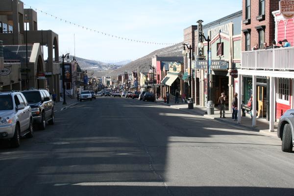 A paved main street lined with multi-story commercial buildings, parked cars, and pedestrians, leading towards a large, tree-covered mountain in the background under a clear sky.