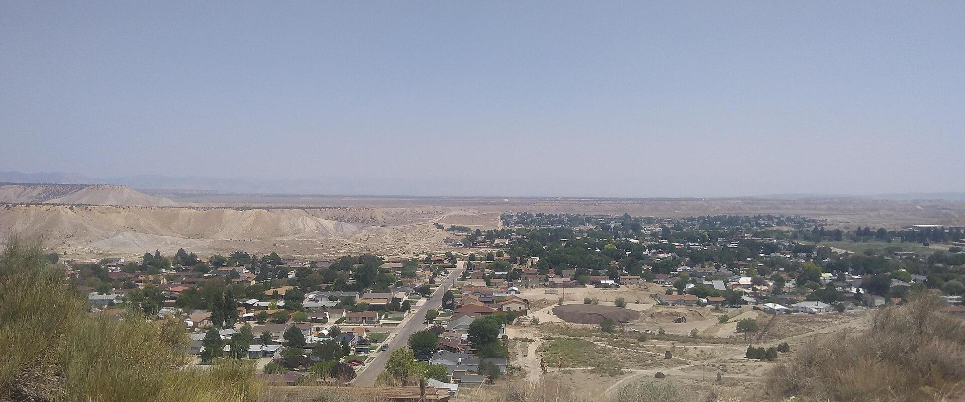 A high-angle, wide shot of a town in a desert valley, looking down from a hillside onto residential streets, houses, and a large, light-colored, excavated area (likely a mine or quarry) in the background.