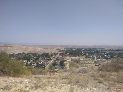 A high-angle, wide shot of a town in a desert valley, looking down from a hillside onto residential streets, houses, and a large, light-colored, excavated area (likely a mine or quarry) in the background.