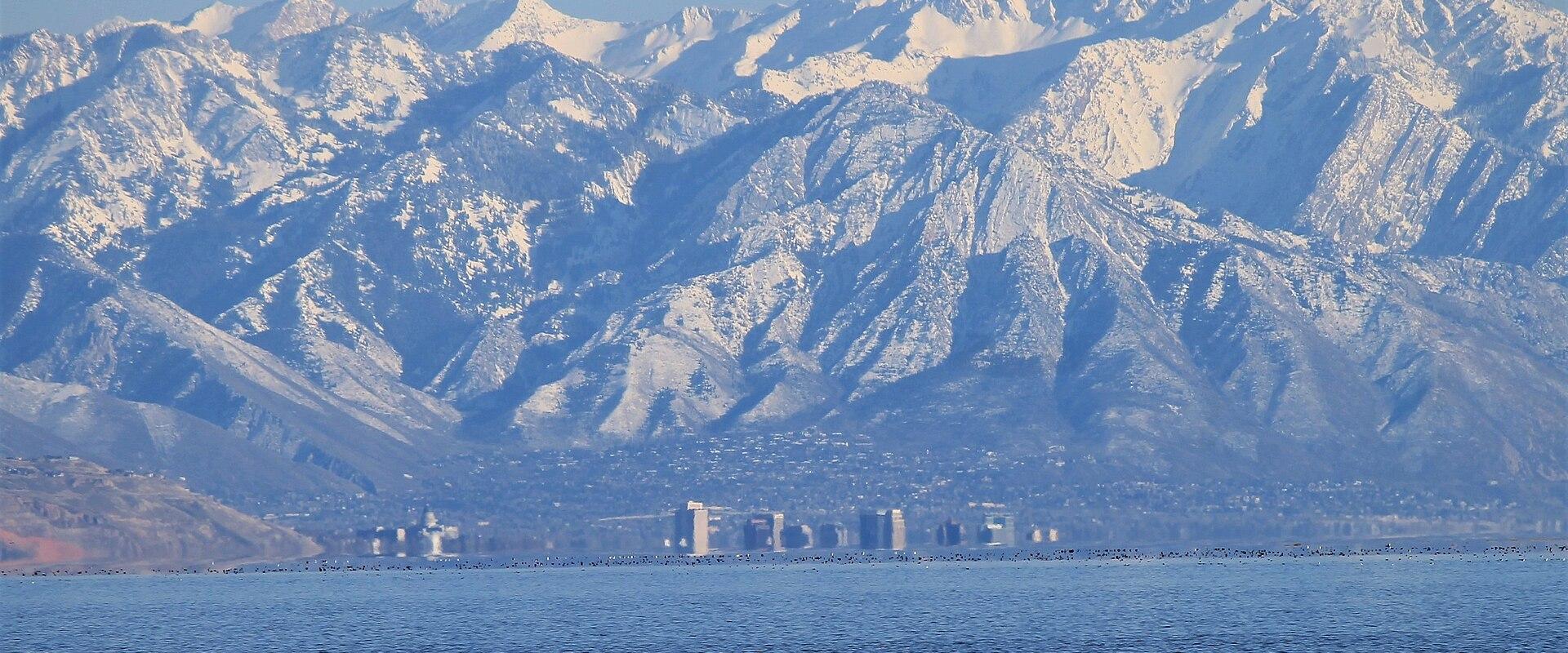 A wide landscape view showing a large body of water in the foreground, a line of buildings and development along the far shore, and a large, snow-dusted mountain range in the background.