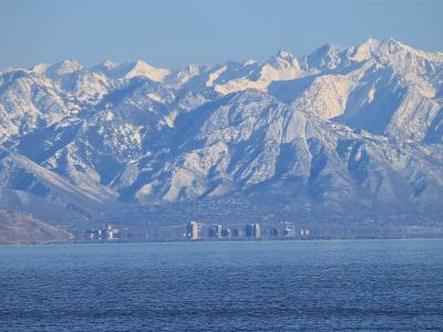 A wide landscape view showing a large body of water in the foreground, a line of buildings and development along the far shore, and a large, snow-dusted mountain range in the background.