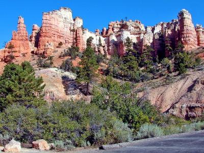 A view of a steep, reddish-orange rock face (hoodoos) with green coniferous trees in the foreground, and a paved road or parking area in the immediate foreground.