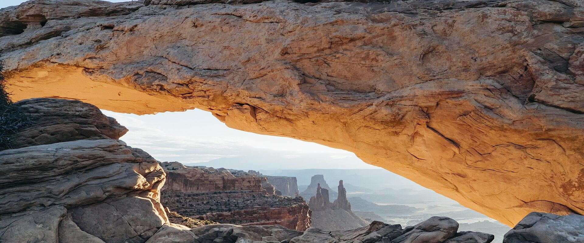 A low-angle photograph of a large, horizontal sandstone arch illuminated by warm sunlight, framing a distant view of canyon mesas and spires under a clear sky.