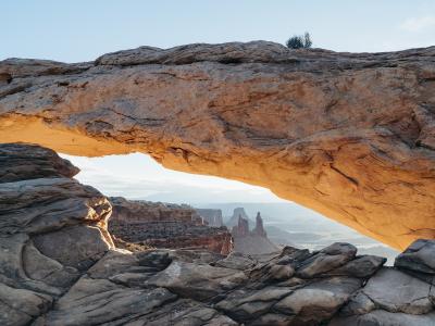 A low-angle photograph of a large, horizontal sandstone arch illuminated by warm sunlight, framing a distant view of canyon mesas and spires under a clear sky.