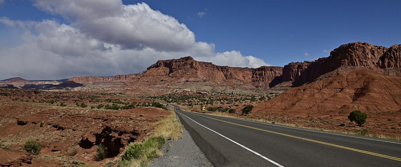 A paved two-lane highway winding through a red rock desert landscape with a prominent, steep cliff face rising on the right side under a blue sky with clouds.