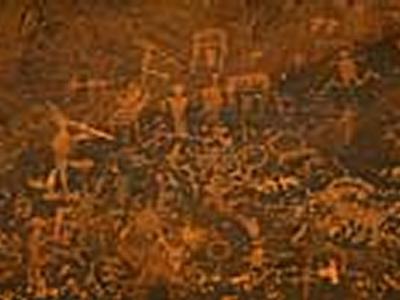 The image displays a close-up, low-light view of a rock surface covered in petroglyphs, featuring stylized anthropomorphic and zoomorphic figures, along with a distinct, large, light-colored geometric shape (possibly a ladder or grid) superimposed or