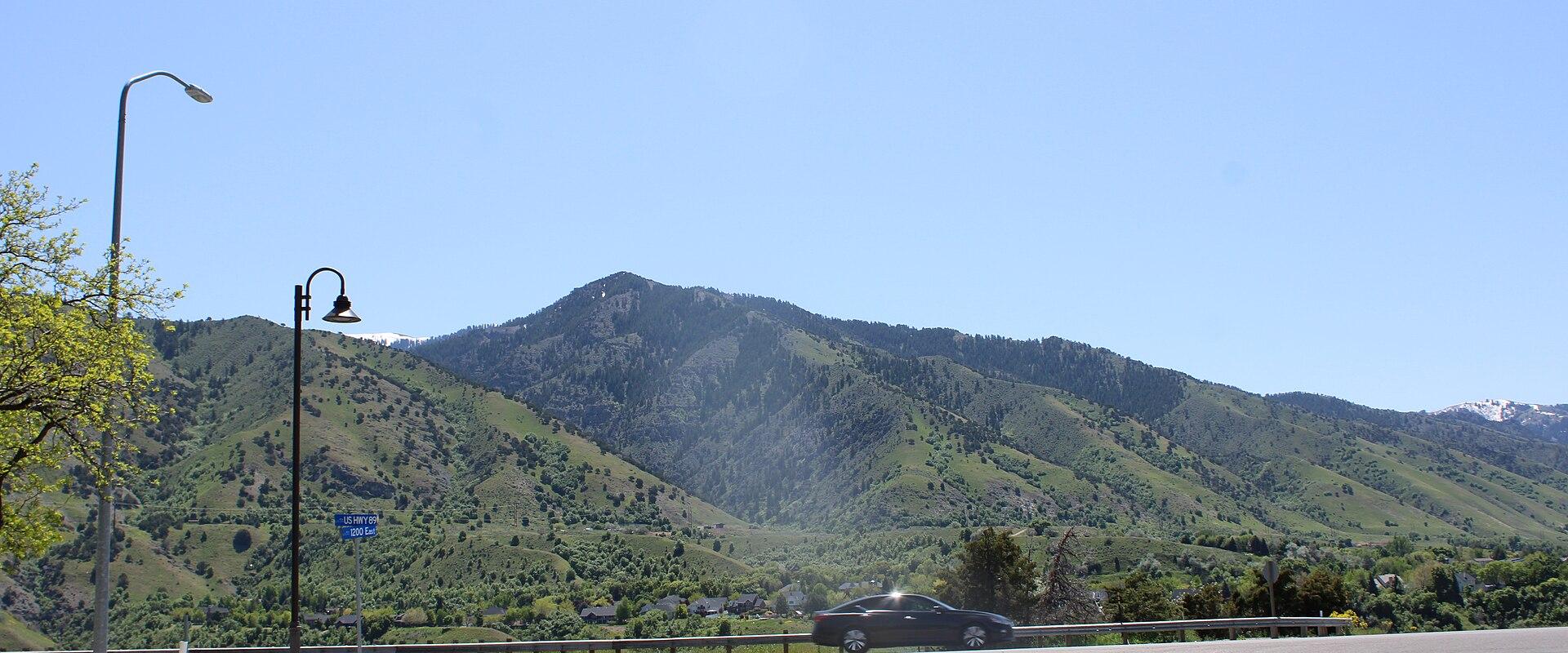 A view of a green, rolling mountain range with a paved road in the foreground, a black car, and a street lamp on the left.
