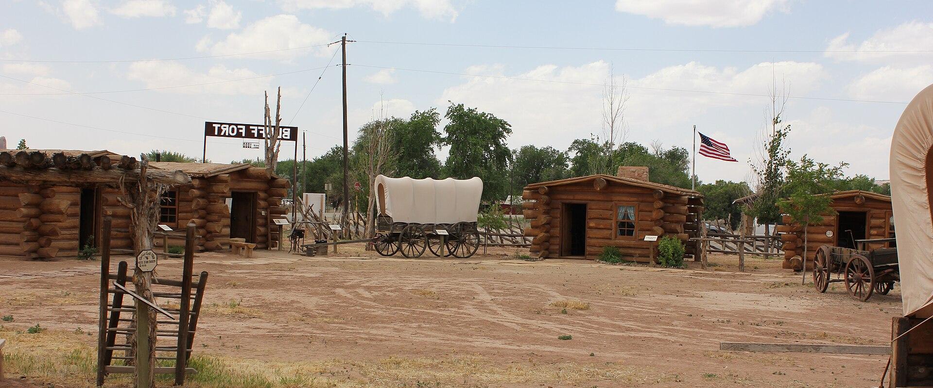 The image shows a collection of historic log structures and covered wagons on a dirt lot, with a large sign in the background that reads "BLUFF FORT" in reverse (indicating the photo is mirrored or taken from the back).