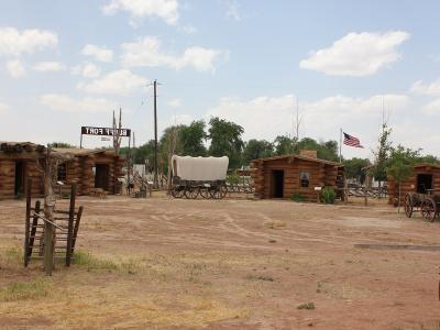The image shows a collection of historic log structures and covered wagons on a dirt lot, with a large sign in the background that reads "BLUFF FORT" in reverse (indicating the photo is mirrored or taken from the back).