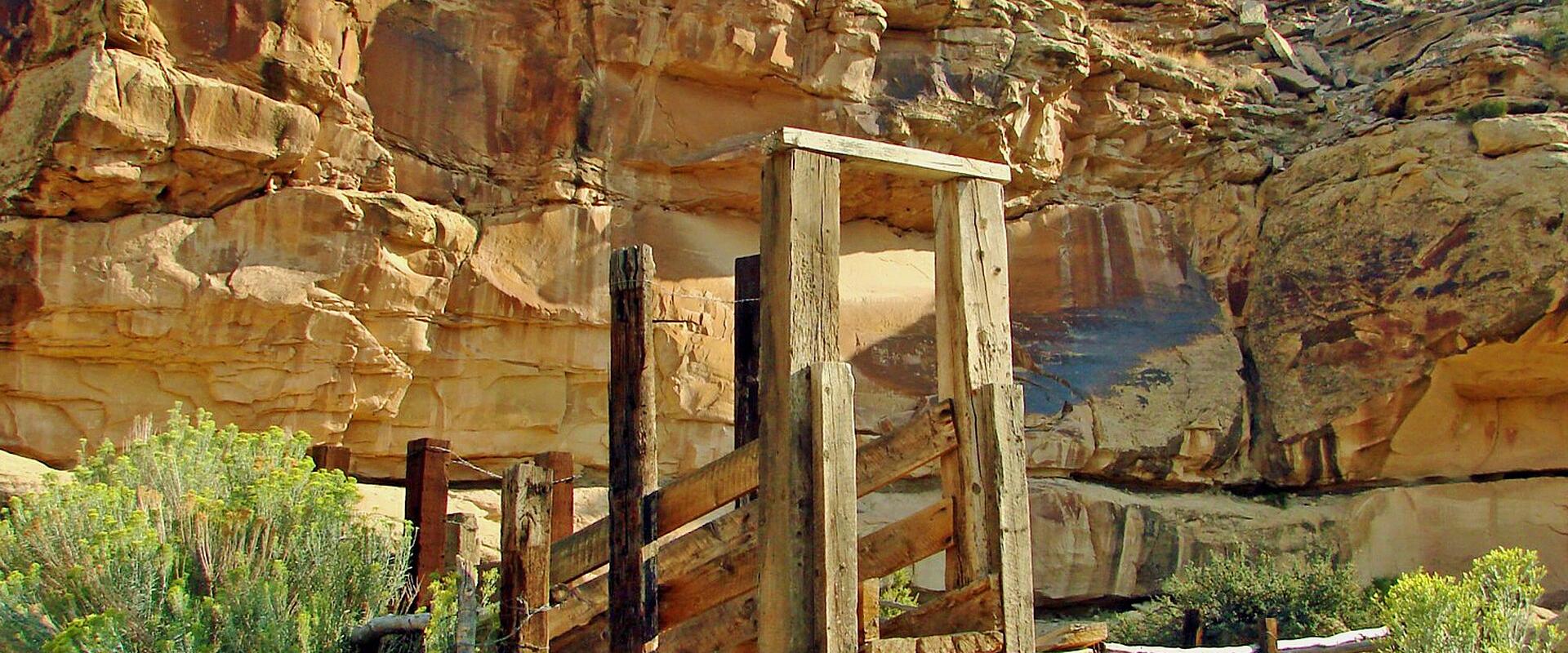 A wooden viewing platform and fence structure built at the base of a sandstone cliff face featuring dark pictographs, with green shrubbery in the foreground.