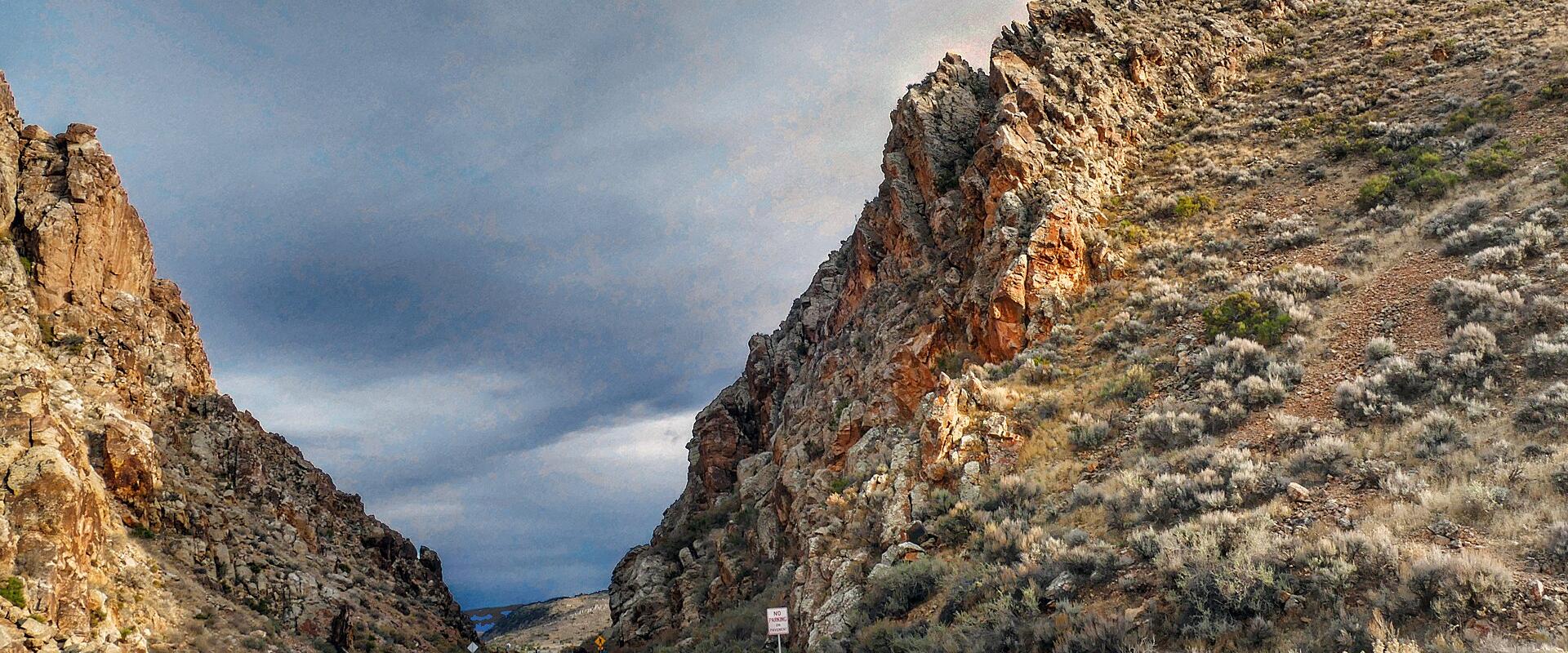 A paved road passing through a narrow, steep-walled rock canyon (the gap) with a person in a red jacket walking on the left shoulder and a white regulatory sign on the right.