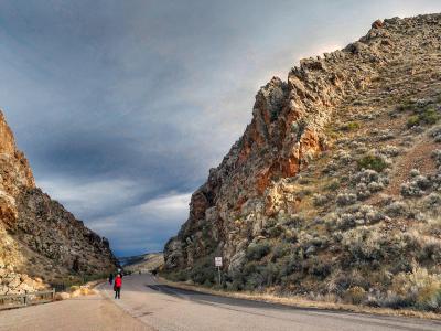 A paved road passing through a narrow, steep-walled rock canyon (the gap) with a person in a red jacket walking on the left shoulder and a white regulatory sign on the right.