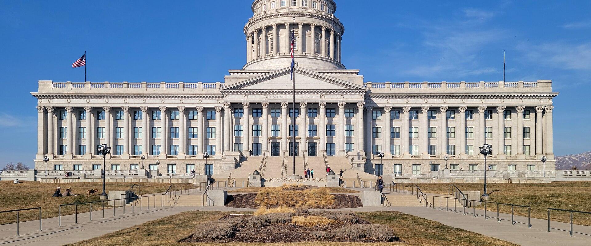 A large, white, neoclassical government building with a prominent central dome, set against a clear blue sky, with a paved forecourt and grassy areas in the foreground.