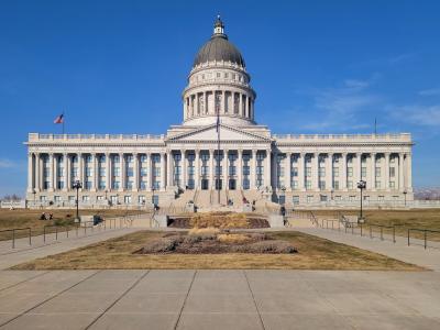 A large, white, neoclassical government building with a prominent central dome, set against a clear blue sky, with a paved forecourt and grassy areas in the foreground.