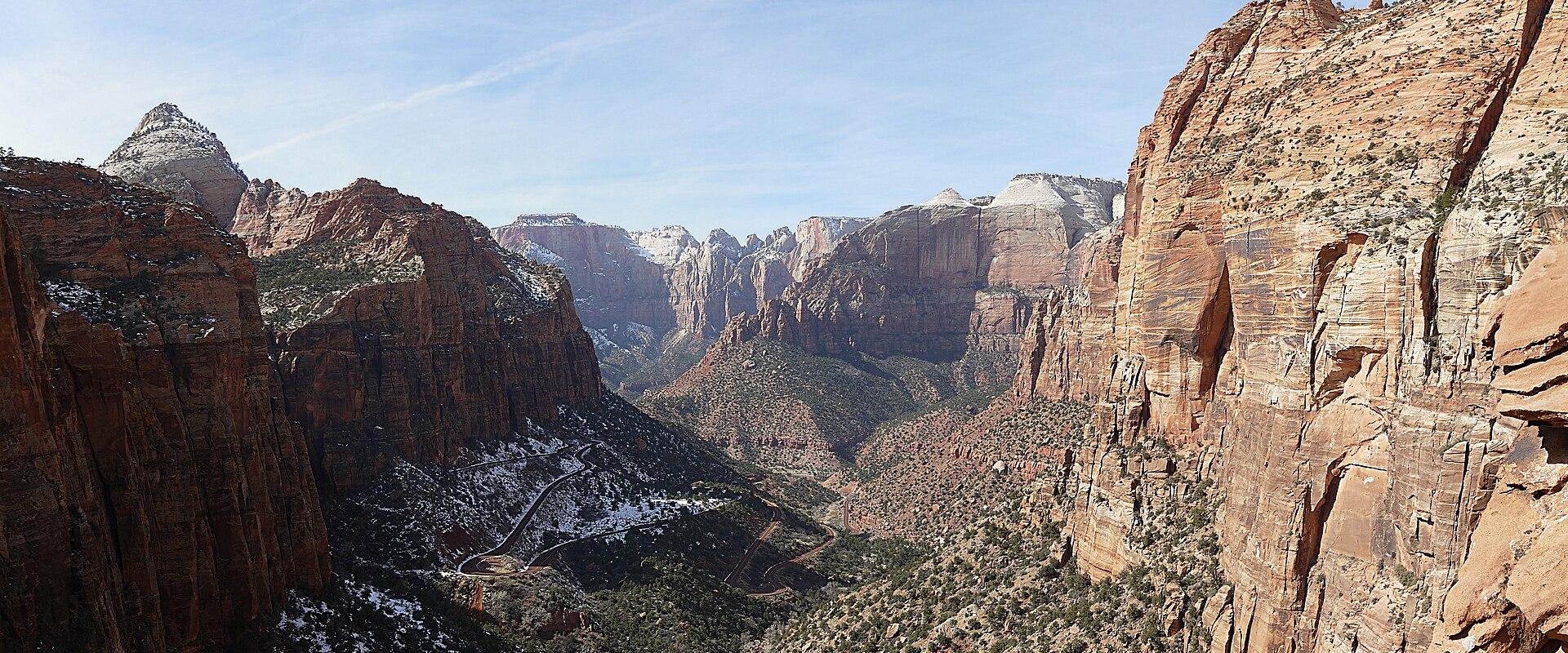 A high-angle, wide landscape view of a deep, narrow canyon with steep, reddish-orange sandstone cliffs, a winding road visible in the valley floor, and a prominent, pointed rock spire (The Watchman) on the left side, all under a blue sky with light c