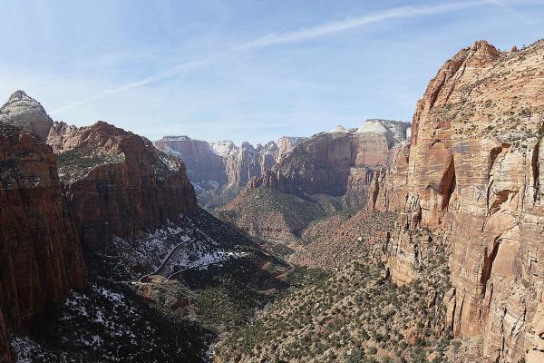 A high-angle, wide landscape view of a deep, narrow canyon with steep, reddish-orange sandstone cliffs, a winding road visible in the valley floor, and a prominent, pointed rock spire (The Watchman) on the left side, all under a blue sky with light c