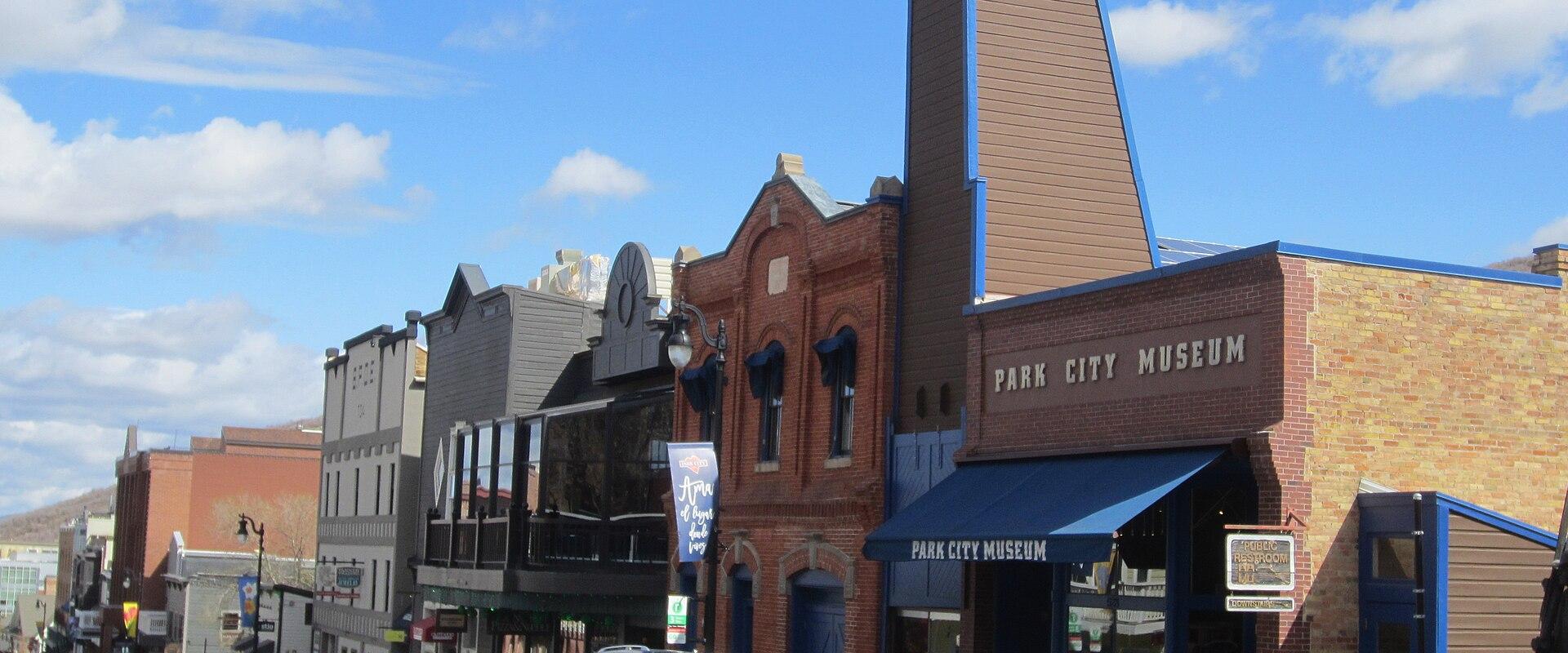 A street-level view of a brick building with a blue awning, identified by large lettering on the facade as the "Park City Museum," situated on a street with other buildings and a parked car.