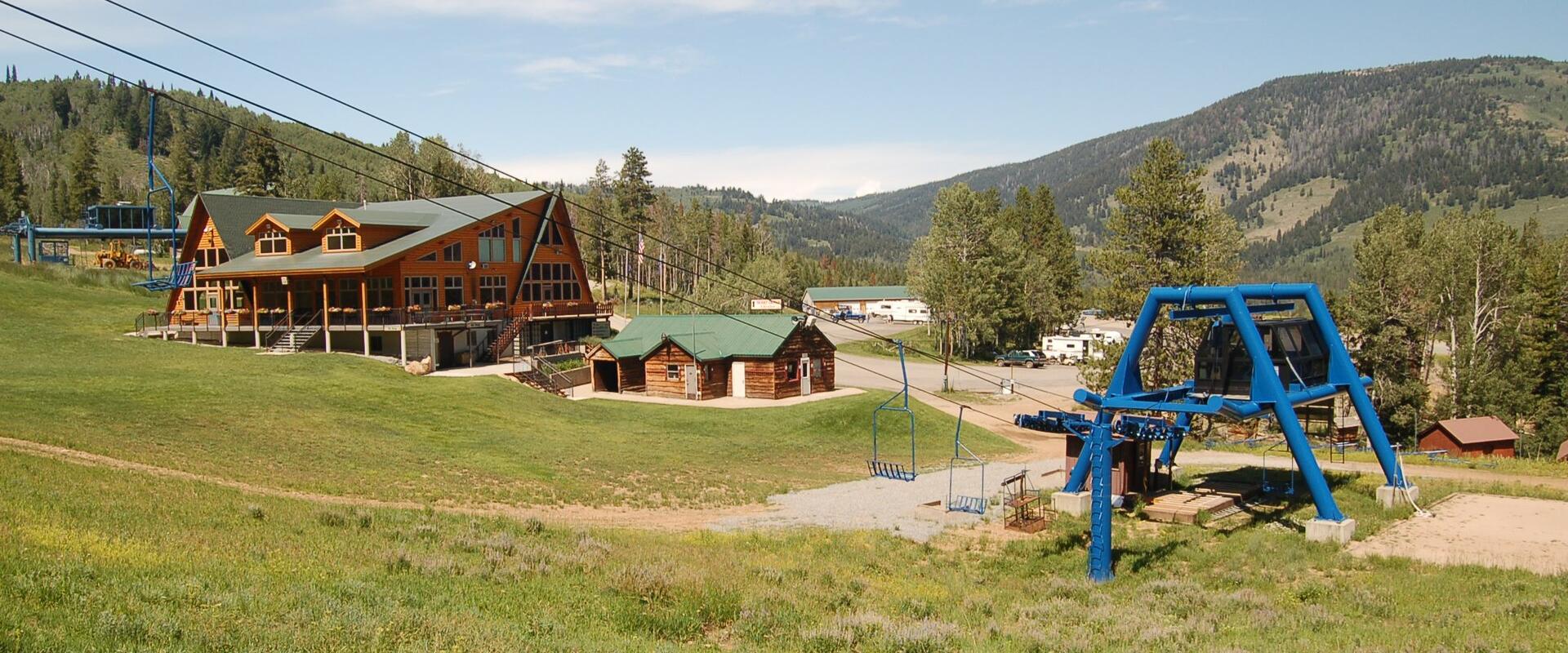 A view of a ski resort base area in summer, featuring a large lodge with a green roof, a smaller wooden cabin, a blue chairlift tower, and a grassy foreground, with forested mountains in the background.